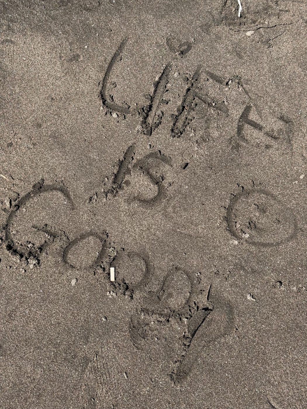 The words life is good and a smiley face written in dark sand